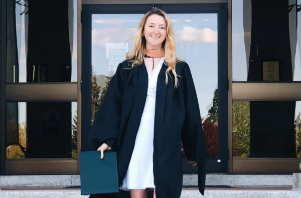 A smiling woman in graduation attire standing in front of the entrance to a building with glass doors.